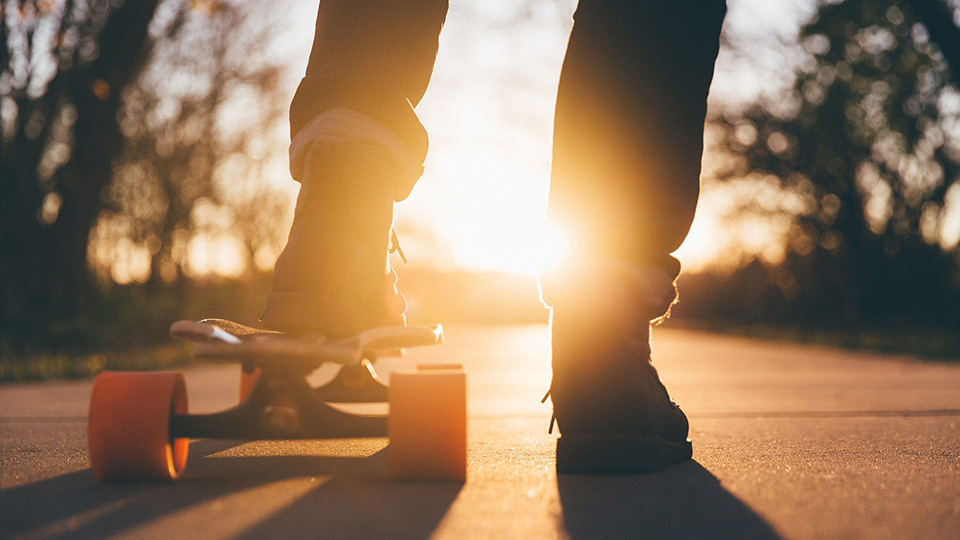 Füße stehen auf einem Skateboard, im Hintergrund ein Sonnenuntergang
